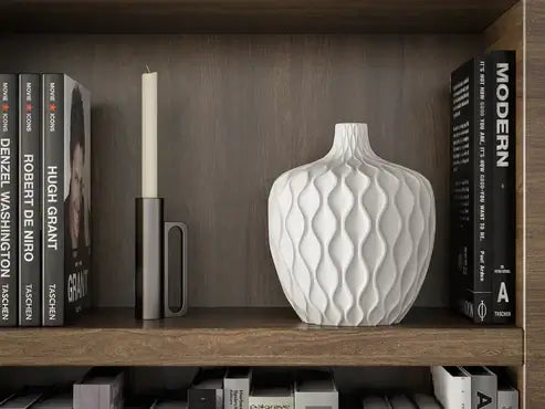 Elegant white wavy vase beside candle and books on wooden shelf, enhancing modern home decor aesthetics.