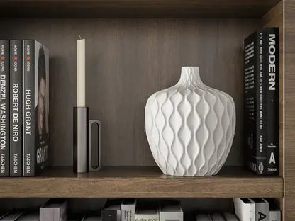 Elegant white wavy vase beside candle and books on wooden shelf, enhancing modern home decor aesthetics.